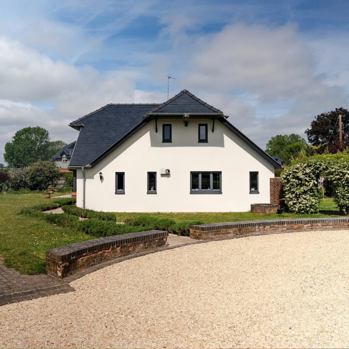 A large white house is in the middle of the image with a curved driveway with new gravel. A pathway connects the driveway to the house. The sky is blue with some clouds and the garden is full of green plants and a large lawn.