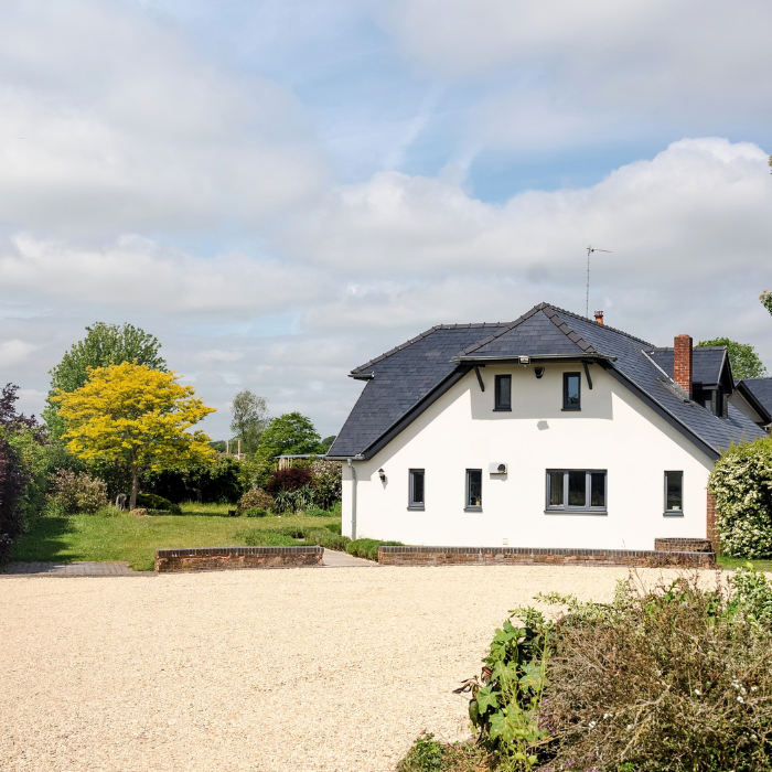 A large white house is in the back of the image with a large driveway with new gravel. There are some bushes in the bottom right corner of the image. The sky is blue with some clouds and the garden is full of green plants and a large lawn.