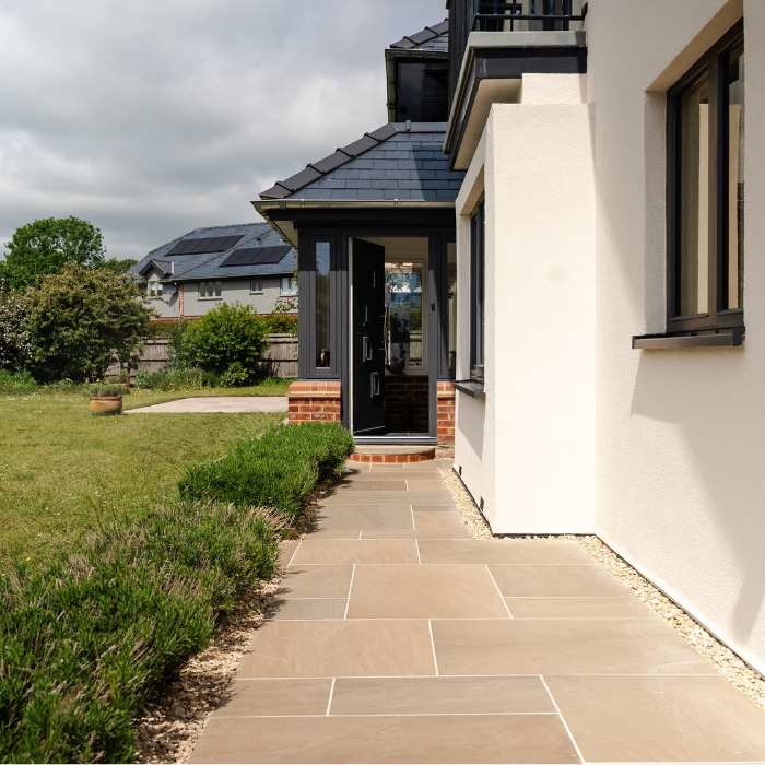 A porcelain pathway lined with small bushes leads to a front door of a house surrounded by a well-maintained garden.